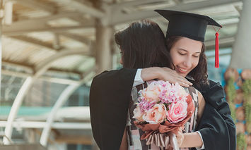 A graduate in a cap and gown hugs another person tightly, holding a bouquet of flowers. They are in a modern, open-air architectural setting with glass and metal structures.