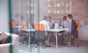 People in business attire, sitting around a conference table with laptops and papers, engaged in discussion within a modern office with glass walls and city buildings visible outside.