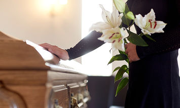 A person in dark clothing holds white lilies and places a hand on a wooden coffin in a softly lit room.